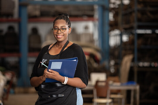 Smiling african american female worker holding blue folder while inspecting car parts warehouse. Concept of automotive maintenance, quality control service. Employees checking inventory.