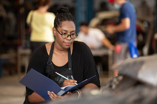 Confident african american female worker holding blue folder while inspecting car parts warehouse. Concept of automotive maintenance, quality control industrial service.