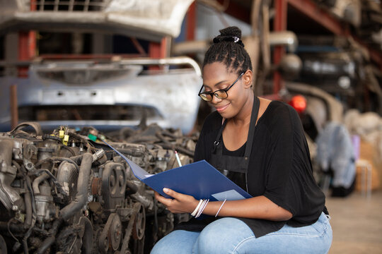Confident african american female worker holding blue folder while inspecting car parts warehouse. Concept of automotive maintenance, quality control industrial service. - Powered by Adobe