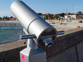 A silver tourist telescope on a waterfront promenade, offering views of the sea and distant town.