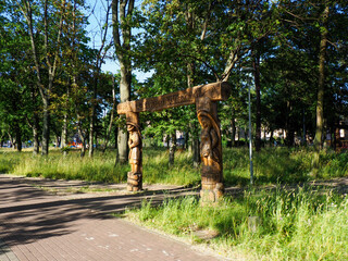 Two elaborately carved wooden posts stand tall beside a gravel path in a sunny park with green...