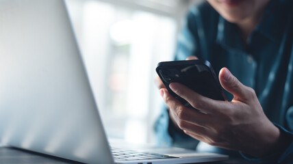 Business woman using smartphone, surfing the internet, searching the information during working on laptop computer at office. Close up, woman using mobile phone for online shopping