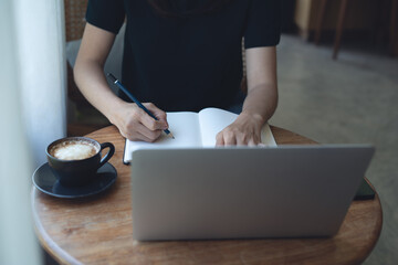 Business woman working on laptop computer and writing on notebook on tablet at coffee shop, remote work. Student studying online and taking note, e-learning, online study concept, closeup