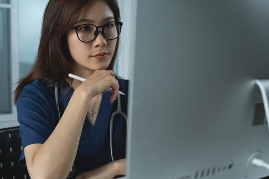 Thoughtful serious asian female doctor sitting at work desk looking at computer screen in office with desktop. Pensive surgeon doctor working on computer, thinking, examining lab result, closeup