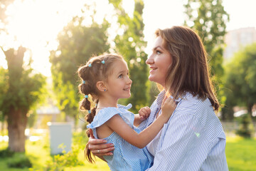 Obraz premium Backlit by the setting sun, a mother hugs her delighted daughter in a striped blue dress, sharing joyful conversation and closeness in a green city park