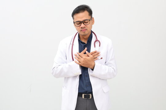 Asian adult male doctor standing wearing doctor's coat, stethoscope hanging around neck, smiling with closed eyes, putting hand on chest, gesture of relief, isolated on white background.
