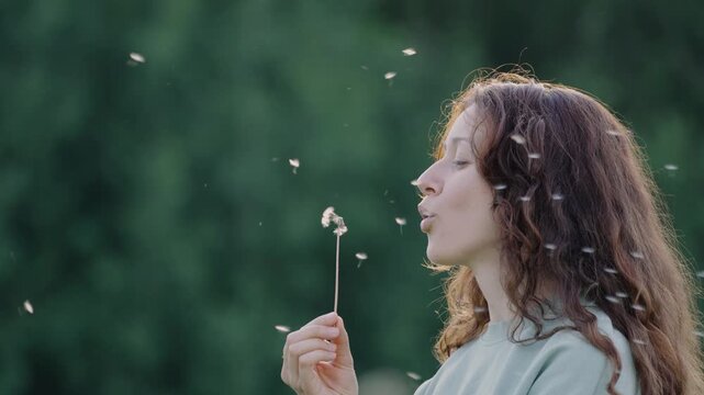 Young woman gently blowing dandelion seeds, creating ethereal patterns against soft green background in dreamy slow-motion sequence highlighting natural beauty and carefree moment