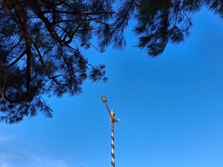 Street lamp pole decorated with ornaments stands tall under a bright blue sky, framed by green tree...