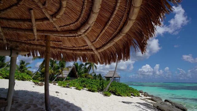 Tropical beach scene with a low-angle view under a thatched umbrella, capturing huts, palm trees, and turquoise waters, ideal for a serene video backdrop.