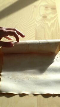 A gentle hand slowly unfurls antique cream parchment paper with deckled edges onto a light wood surface, highlighted by soft natural light and macro detail in a vertical video. Serene concept of