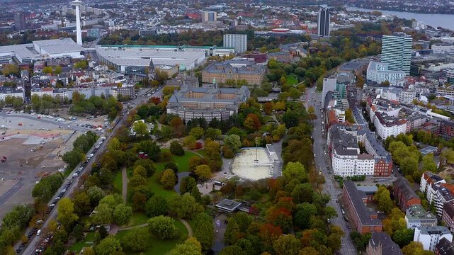 4K drone panorama of Hamburg: Planten un Blomen park in autumn, the Museum of Hamburg History and Messe halls beneath the Heinrich-Hertz-Turm, with the Alster skyline beyond.