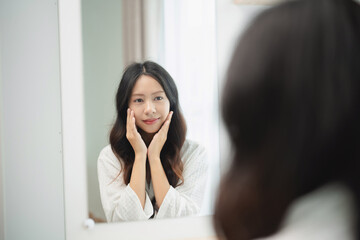 Young woman in white robe admiring her reflection in a mirror while touching her face, showcasing self-care and beauty routine in bright room