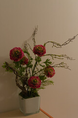 A white vase holds a floral arrangement of artificial red ranunculus flowers, green leaves, and...