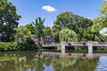Obraz premium A picturesque canal scene in Gouda, Netherlands, featuring a charming bridge adorned with flowers, surrounded by lush green trees and traditional Dutch buildings under a clear blue sky.