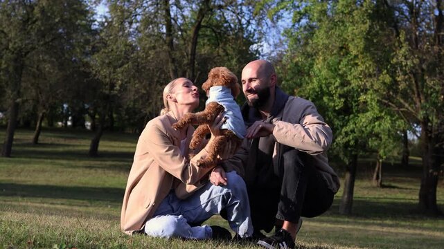 Toy poodle being lovingly held by a woman while a man crouches nearby in a vibrant autumn park, showcasing the joy of pet companionship in nature