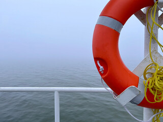 Orange lifebuoy with yellow rope attached to a ship railing against a foggy, calm sea background.