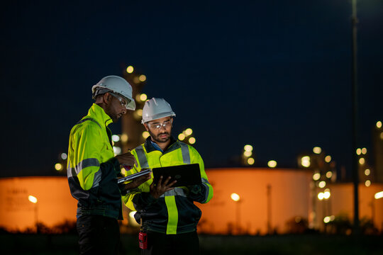 Engineers wearing safety gear, including hard hats examining survey are using tablet collaborating and discussing on screen work plant site use with night lights of oil refinery industry background