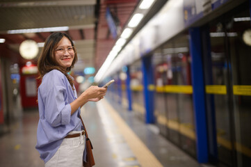 A smiling woman stands at a subway station, holding a smartphone, with empty platforms and clocks...