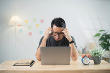 Young man feeling stressed while working at home on a laptop, struggling to focus and under...