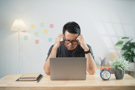 Young man feeling stressed while working at home on a laptop, struggling to focus and under pressure in a bright and organized workspace.
