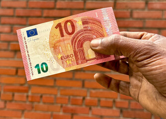 A man holds a 10 Euro banknotes in his hand, over plain brick background. A black man holds 10...