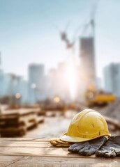 Obraz na płótnie Canvas Yellow hard hat and protective gloves lying on a wooden plank at construction site with crane and excavator in the background