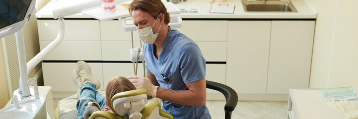 Caucasian male dentist examining teeth of Caucasian child patient in dental chair, both wearing...