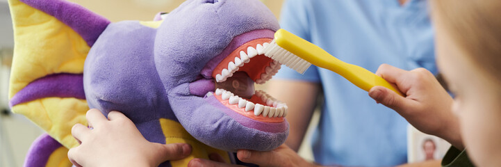Caucasian child holding large toothbrush cleaning teeth of purple plush toy dragon while adult...