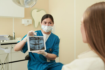 Young adult Caucasian female dentist wearing medical mask, showing dental X-ray image on digital...