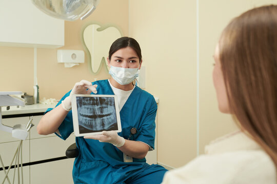 Young adult Caucasian female dentist wearing medical mask, showing dental X-ray image on digital tablet to young adult Caucasian woman patient during consultation in dental clinic