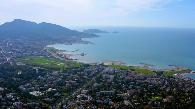 4K drone panorama of Marseille&rsquo;s Prado shoreline : turquoise Mediterranean, curving bays and breakwaters, Borely Hippodrome racetrack and coastal suburbs beneath rugged Provencal mountains.