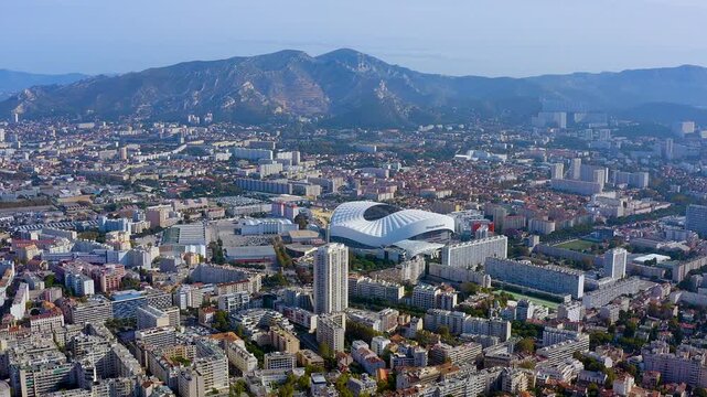 4K drone panorama of Marseille : the Orange Velodrome gleams amid dense city blocks, framed by Provencal mountains and Mediterranean light - epic urban skyline and sports landmark.