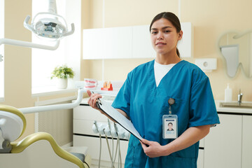 Portrait of young adult woman standing in dental clinic holding clipboard wearing medical scrubs...
