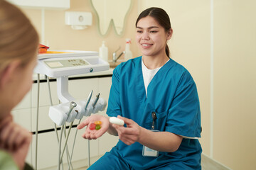 Young adult woman dentist showing dental hygiene tools and colorful dental models to patient in...