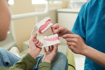Caucasian child sitting while young adult Caucasian man in medical uniform demonstrating dental hygiene using large plastic teeth model and toothbrush in dental clinic