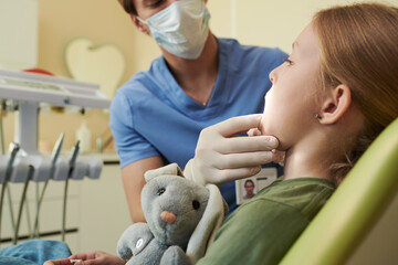 Caucasian girl sitting in dental chair holding stuffed animal, while young adult Caucasian male...