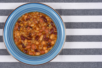 A portion of bean soup with various vegetables and legumes is in a blue, striped bowl, photographed from above on a striped tablecloth