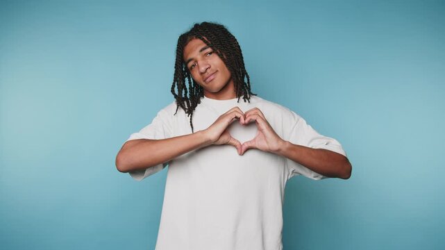 footage of young man expresses love and positivity by forming a heart shape with his hands on light blue background