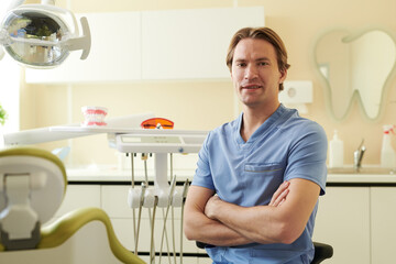 Portrait of young adult Caucasian man dentist sitting with arms crossed in modern dental clinic,...