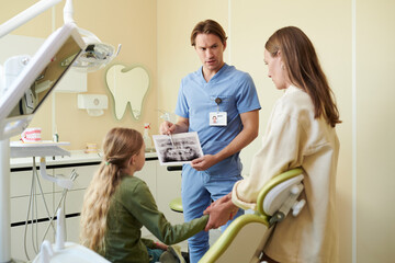 Caucasian male dentist showing dental X-ray to Caucasian teenage girl sitting in dental chair while Caucasian woman, likely mother, standing beside holding teenagers hand