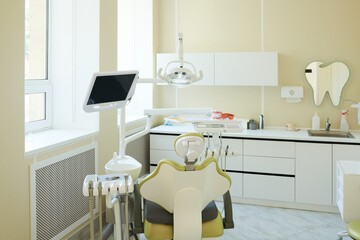 Modern dental office showing empty dental chair with equipment and monitor positioned near window, clean workspace featuring organized cabinets and dental tools on countertop