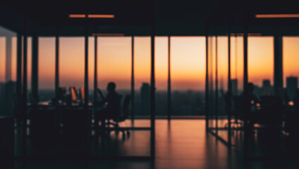 Silhouetted people working collaboratively in an office space featuring large windows with a warm, vibrant sunset view casting natural light and long shadows inside.