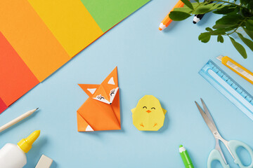 Origami fox and chick surrounded by colorful paper and craft tools on a blue background. Concept of handmade art, creativity, and design.