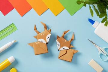 Two origami deer with colorful paper and craft tools on a blue background. Handmade art, creativity, and design for kids.