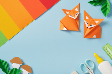 Two origami foxes with mushrooms and leaves surrounded by colorful paper and craft tools on a blue background with copyspace.