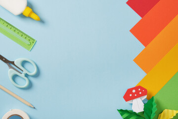 Origami mushroom and leaves with colorful paper and craft tools on a blue background. Idea of handmade art, creativity, and design. Copyspace.