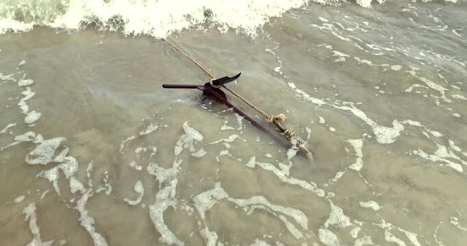 In slow motion, foamy water drifts over a rusty grapnel anchor, tied with a thick rope and submerged in the shallows of a sandy beach.