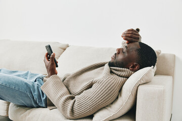 A thoughtful African American man relaxing on a beige couch while using a smartphone, indicating a moment of introspection and connection with technology