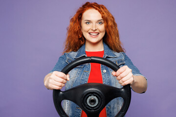 Young smiling happy fun ginger woman she wears red t-shirt denim shirt hold steering wheel driving car look camera isolated on plain pastel light purple background studio portrait. Lifestyle concept.