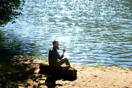 Bearded man sitting on log by lake drinking coffee on sunny day. Outdoor relaxation and travel lifestyle concept - Powered by Adobe